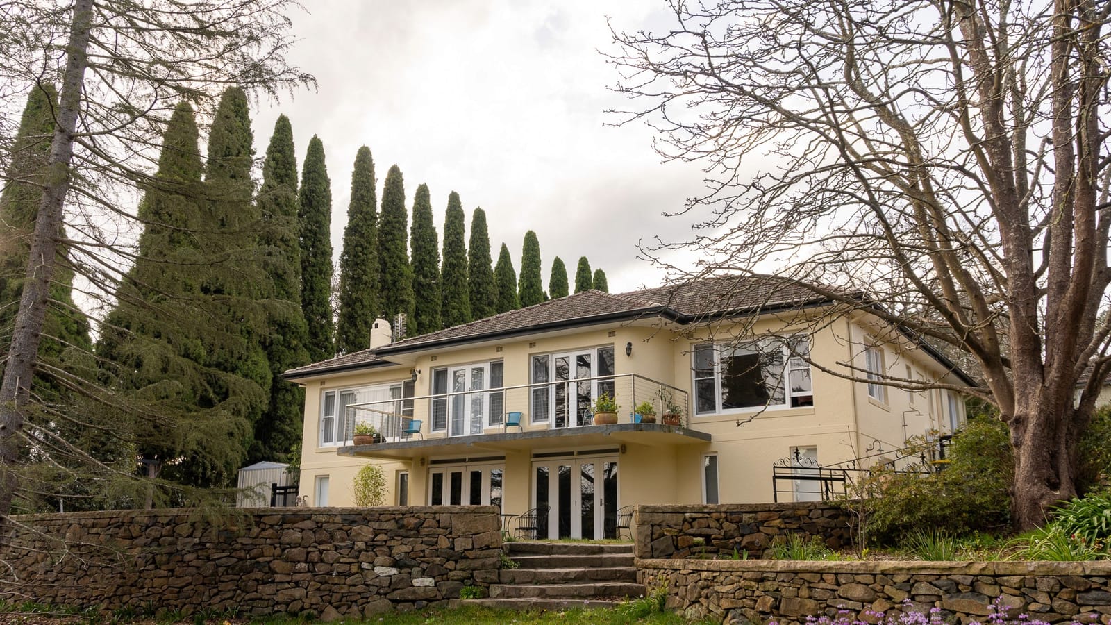 A heritage house standing behind a stone wall, framed by tall cypress and bare winter trees.